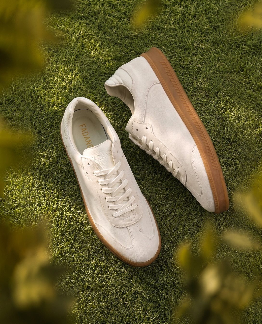 Cream suede sneakers with tan soles resting on a lush green grass lawn.