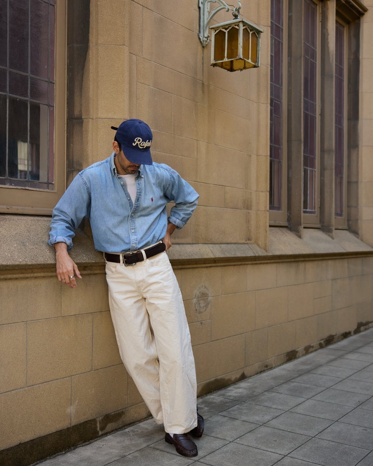 Man in a light blue denim shirt and white trousers leaning against a stone wall.