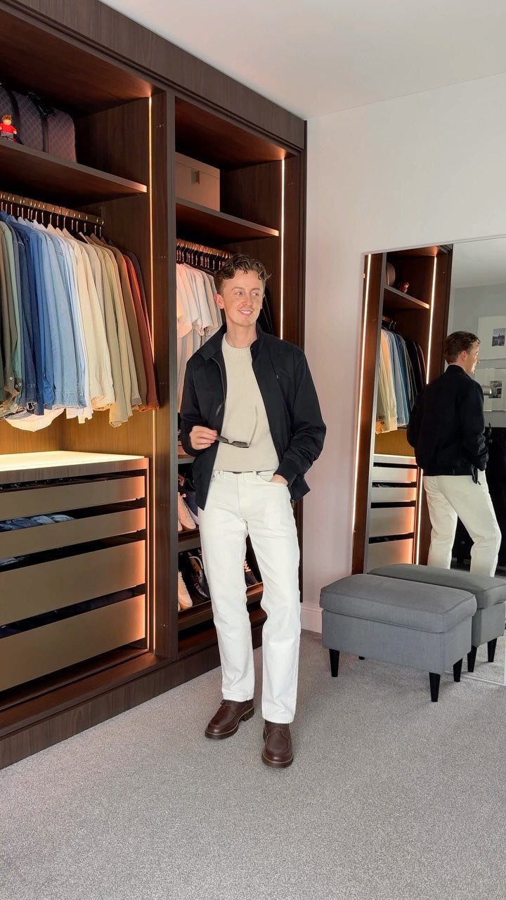A smiling man wearing a black jacket over a cream sweater and white pants in a closet.