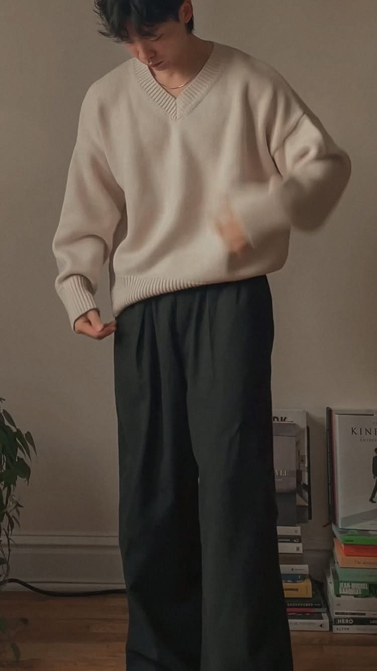 Man wearing an oversized cream V-neck sweater and wide-leg black trousers standing indoors.