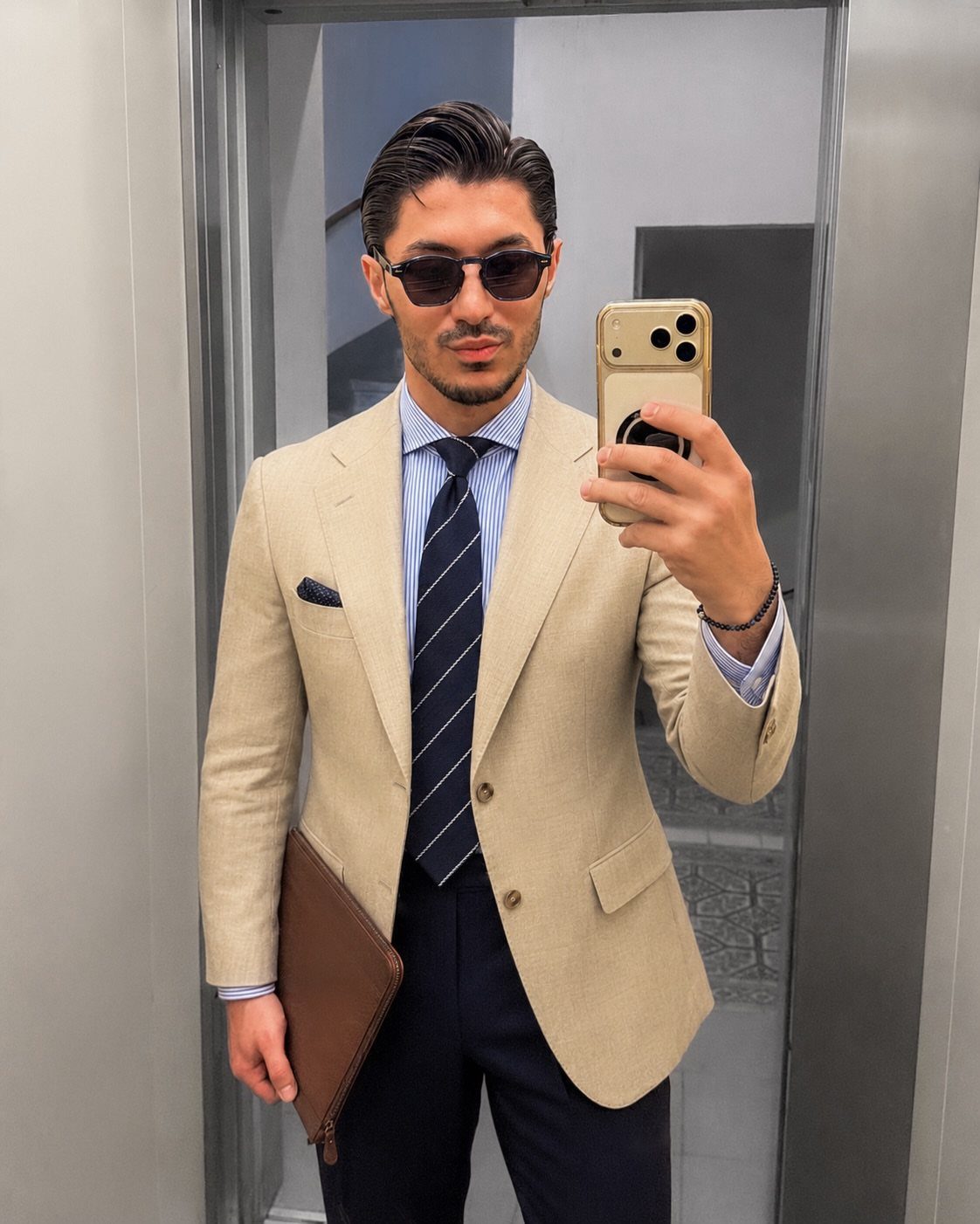 Man in a beige blazer, striped tie, and sunglasses taking a mirror selfie.