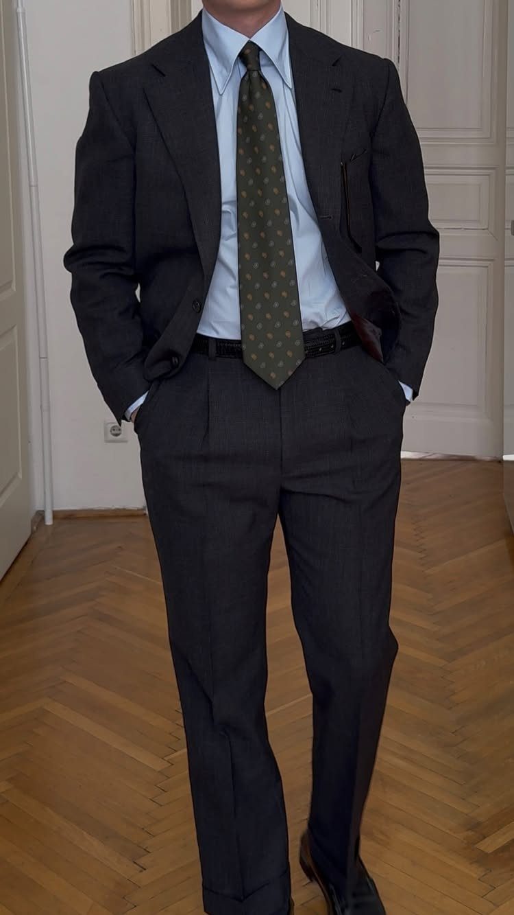 A man wearing a dark charcoal grey suit with a light blue shirt and green tie.