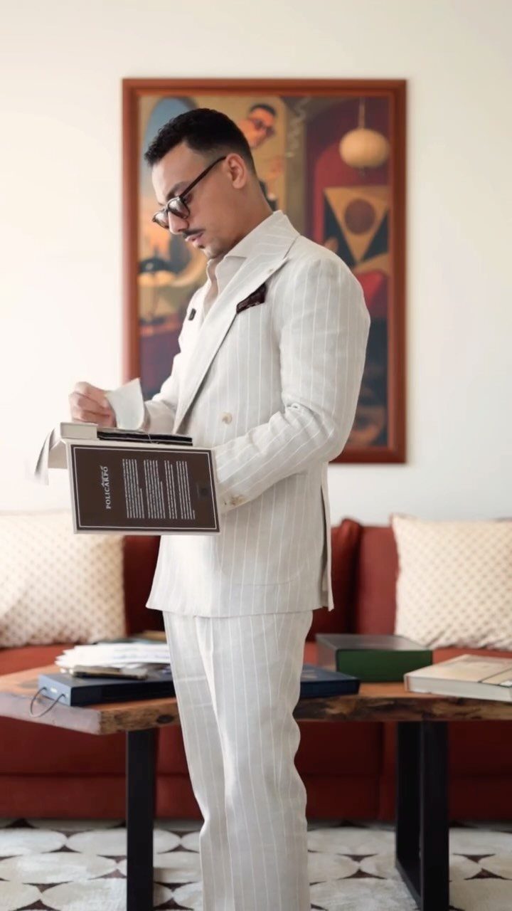 A man in a light-colored striped linen suit stands in a living room holding a book.
