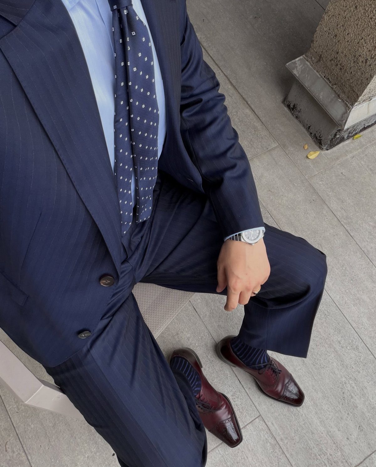 Man in a navy pinstripe suit, polka dot tie, and burgundy loafers sitting outdoors.