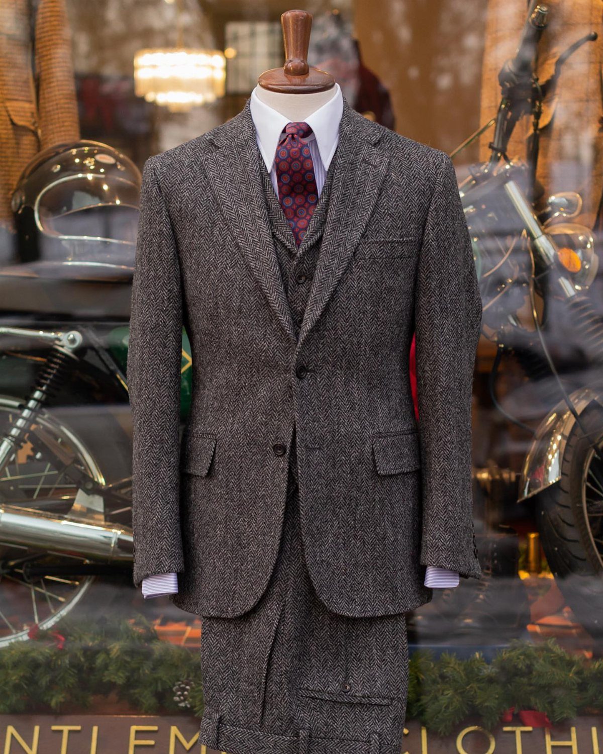 A grey tweed three-piece suit with a burgundy tie displayed on a mannequin.
