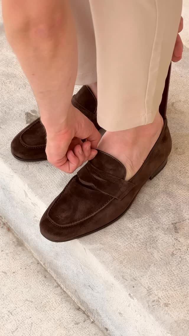 A person putting on dark brown suede loafers while wearing beige trousers.