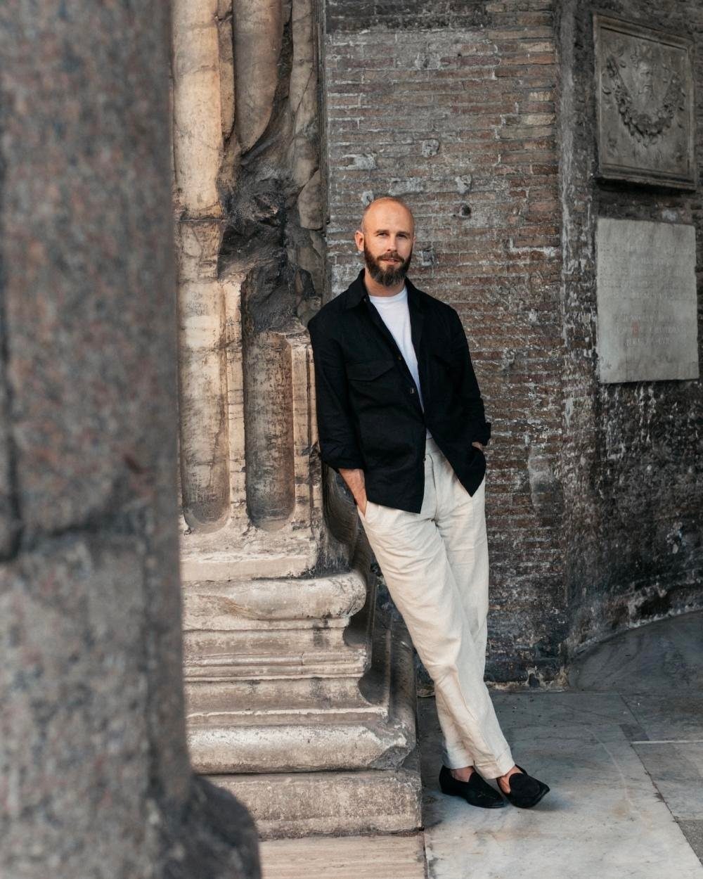 Man in a black shirt and light trousers leaning against a stone pillar outdoors.