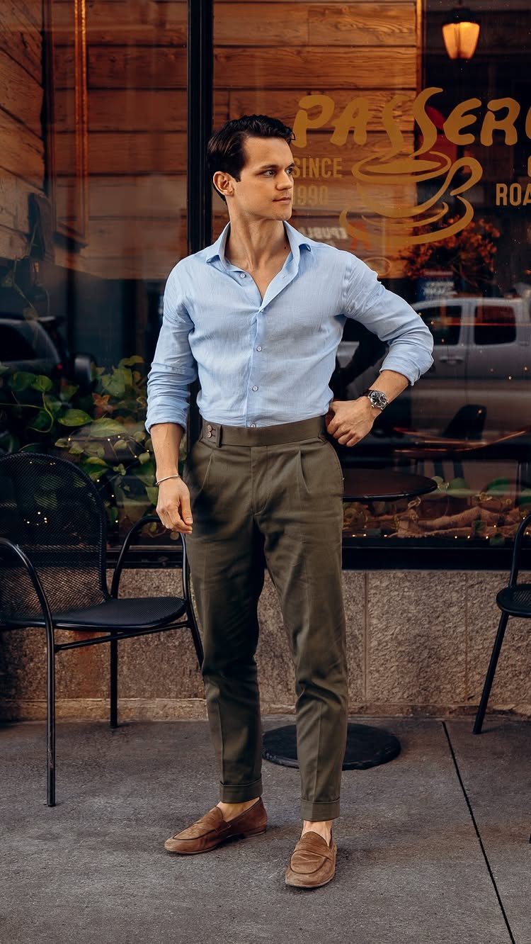 Man in light blue shirt and olive green trousers posing outdoors near a cafe window.
