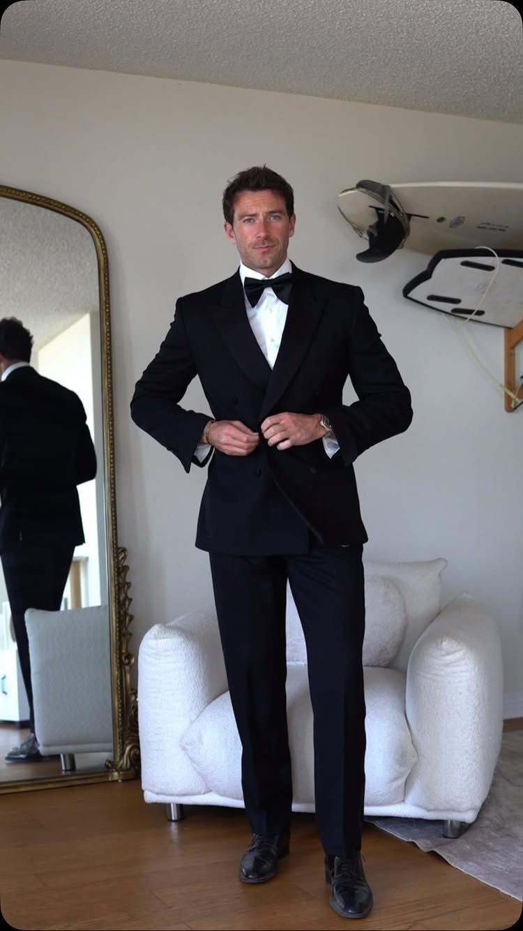 Man in a black tuxedo and bowtie standing in a room adjusting his jacket.