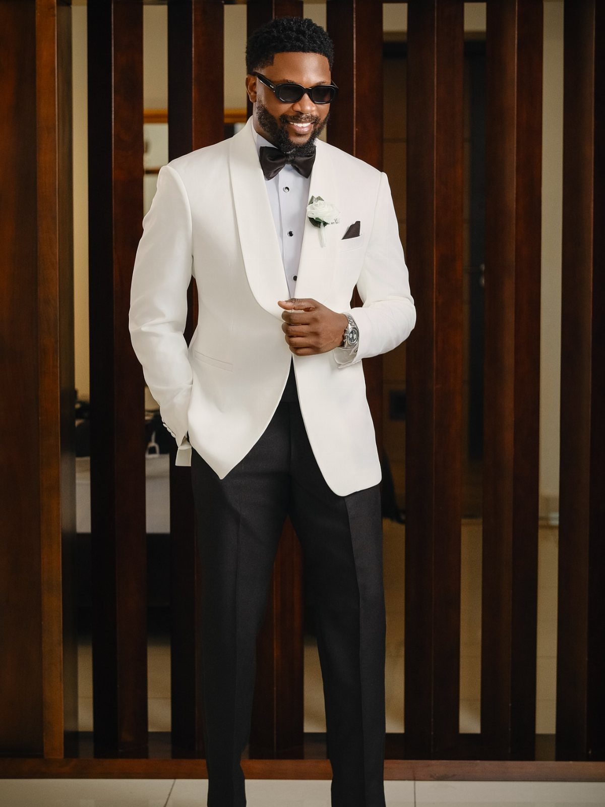 A man in a white tuxedo jacket and black bow tie smiling against wooden slats.