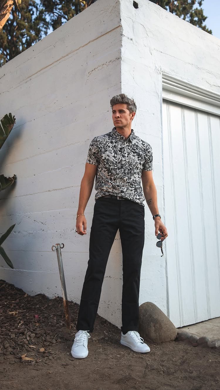 Man in a patterned short-sleeved shirt, black pants, and white sneakers standing by a white building.