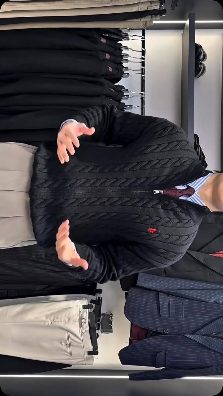 Man in a black cable-knit sweater and tie standing in a clothing store.