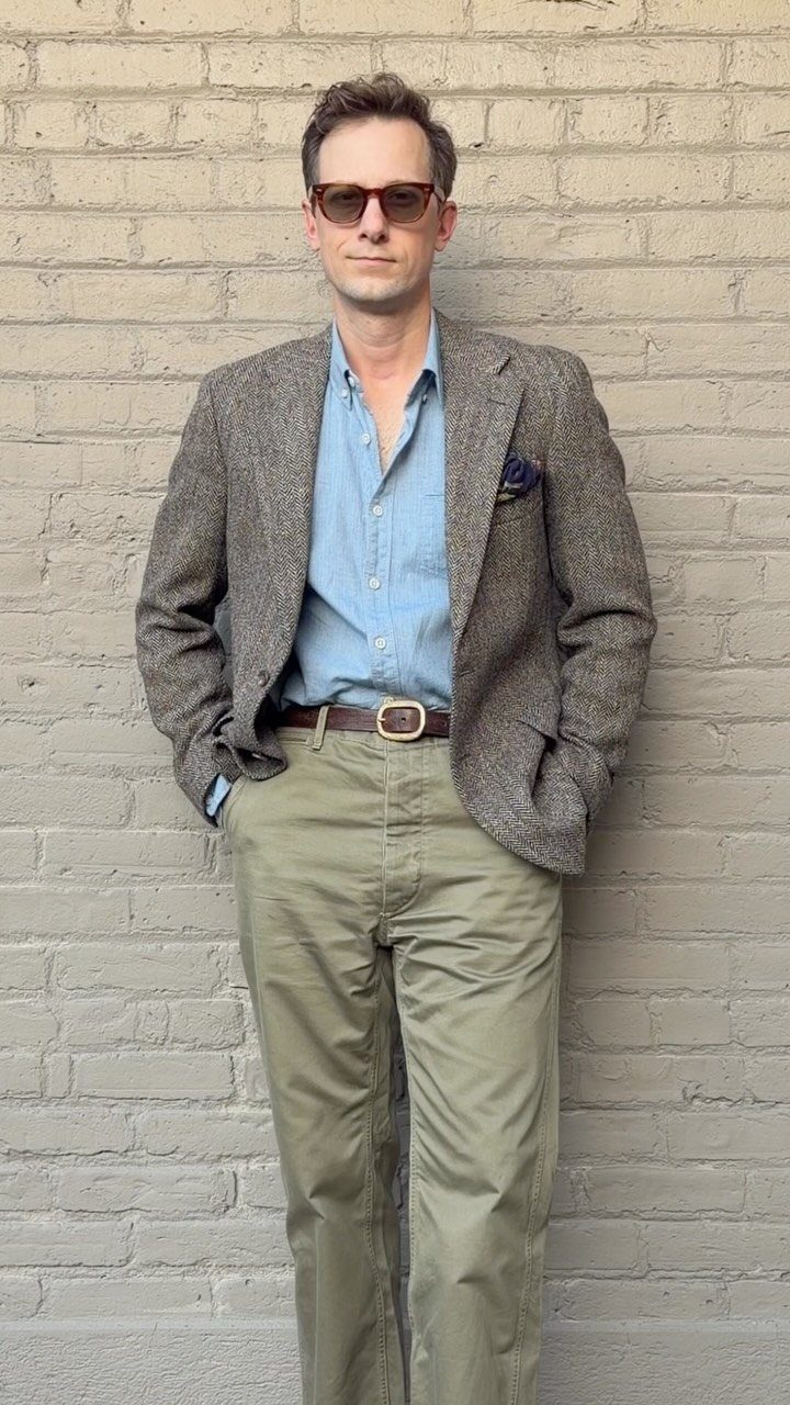 Man in a grey blazer, blue shirt, and olive trousers standing against a brick wall.