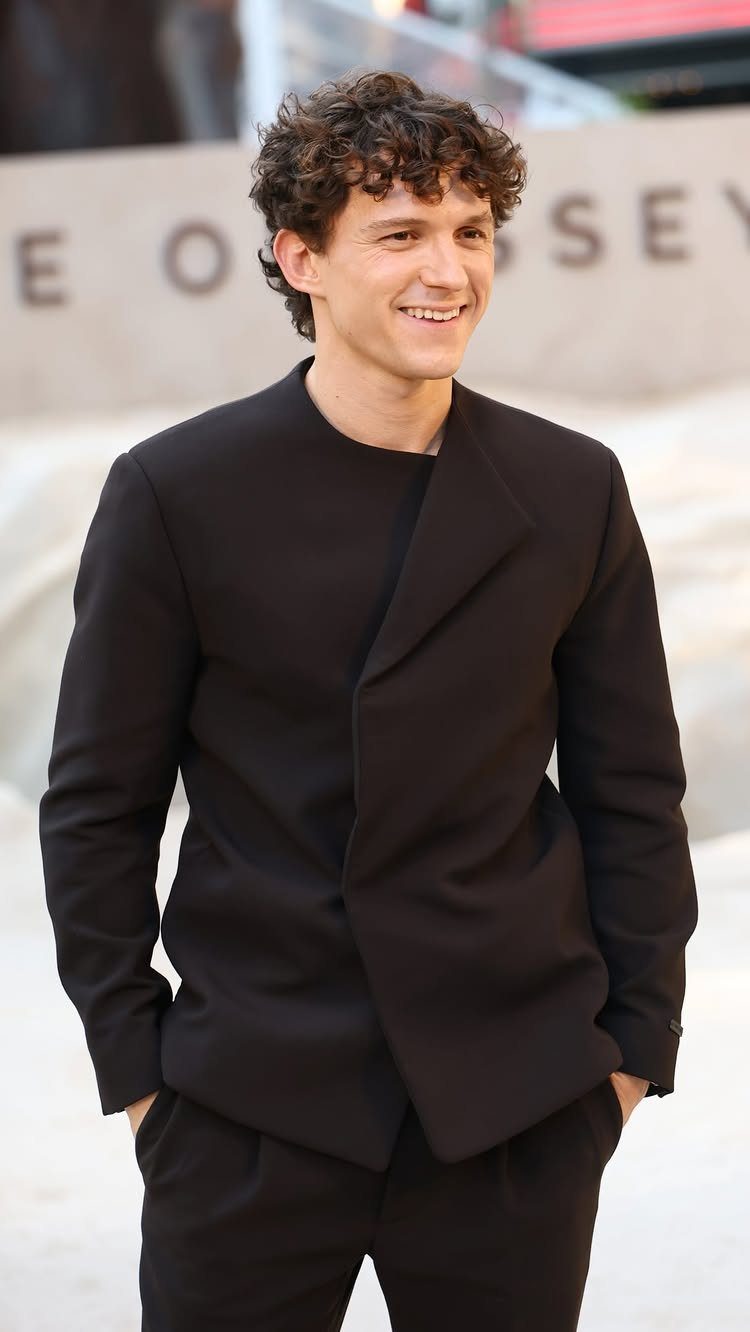 A smiling man with curly hair wearing a minimalist black wrap-style jacket and matching trousers.