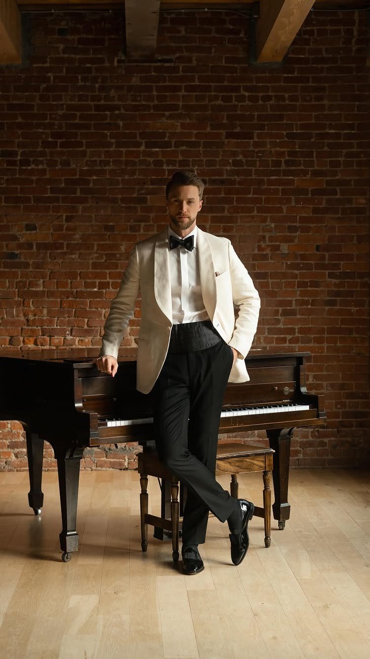 Man in a white tuxedo jacket and black bow tie leaning against a grand piano.