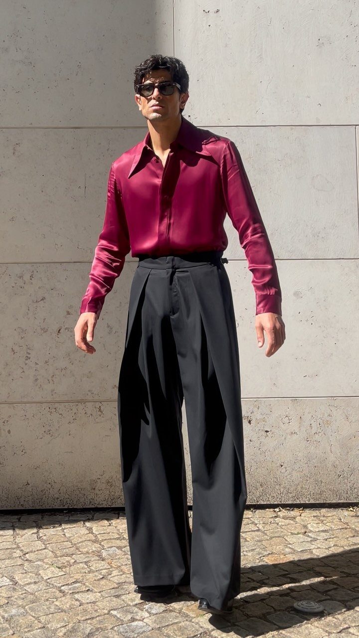 A man in a maroon silk shirt and wide-leg black trousers posing against a stone wall.