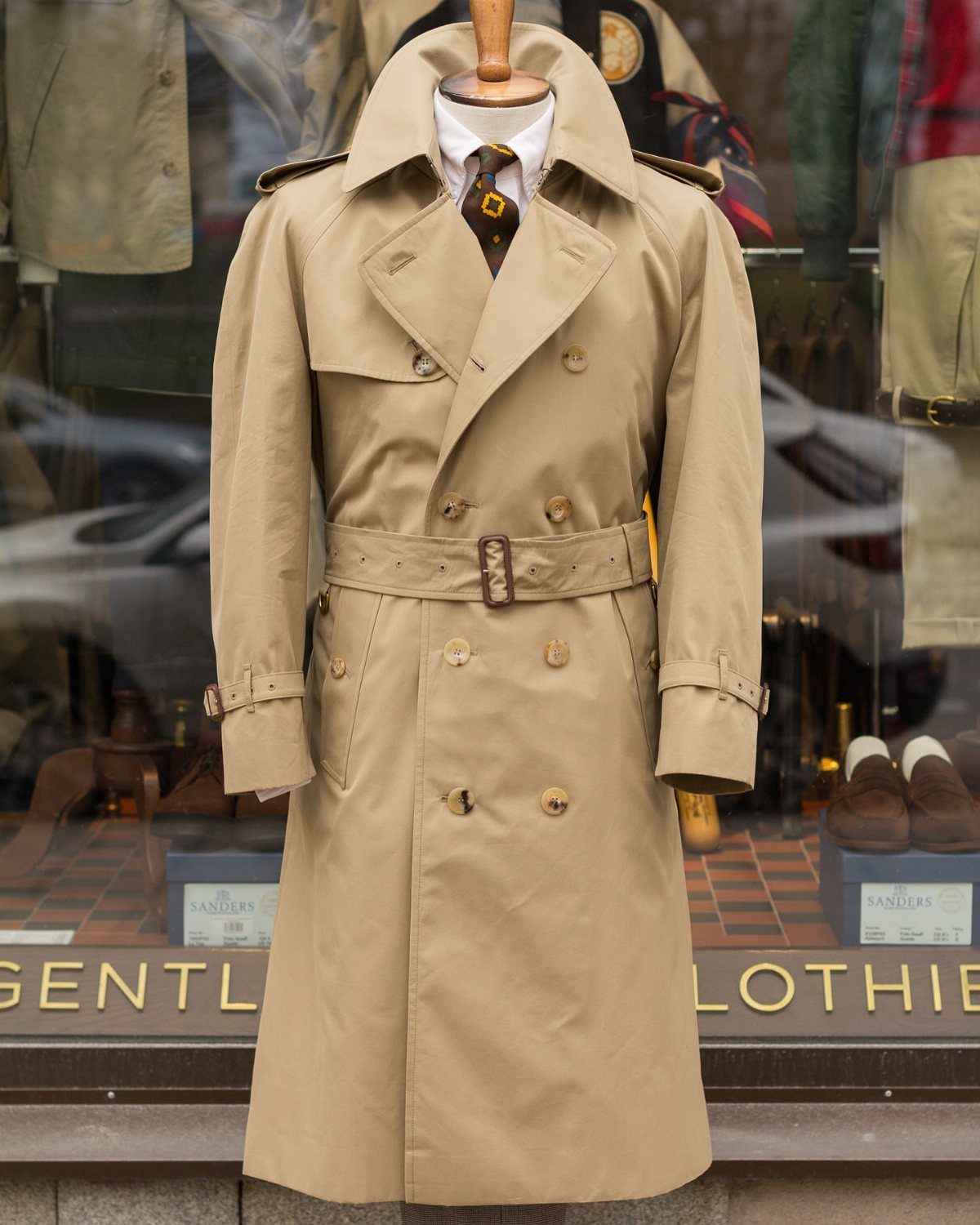 A beige trench coat on a mannequin wearing a white shirt and tie in a shop window.