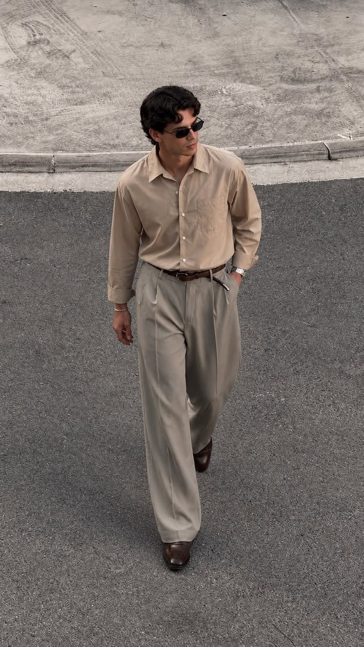 A man in tan clothing and pleated dress pants walking on an asphalt street.