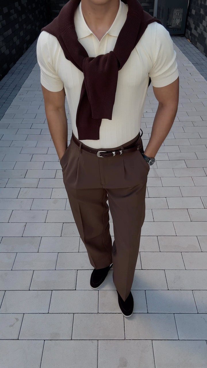 Man in cream polo, brown pleated trousers, and a dark sweater draped over his shoulders.