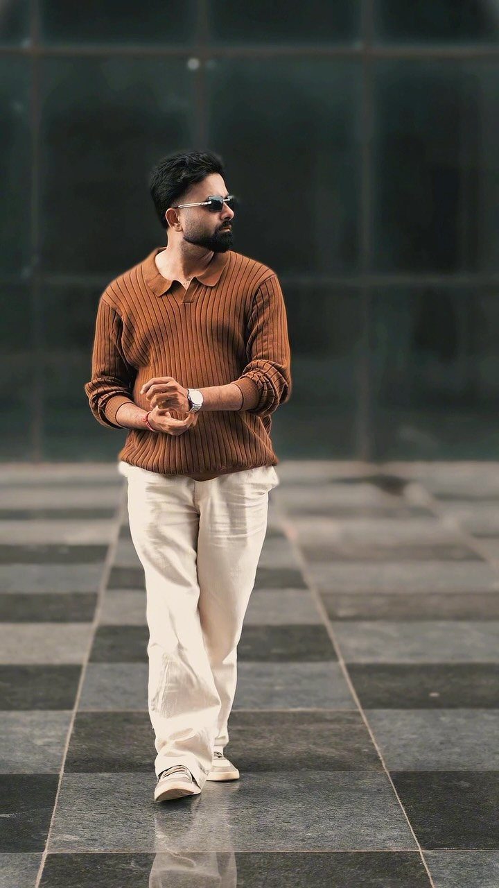 A man in a brown textured polo and cream trousers posing outdoors.