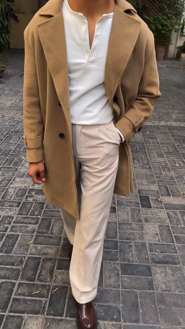 Man wearing a tan overcoat, white henley shirt, and beige trousers on a cobblestone path.