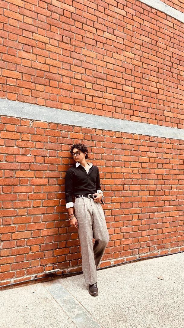 A man in a black polo and grey trousers leaning against a red brick wall.