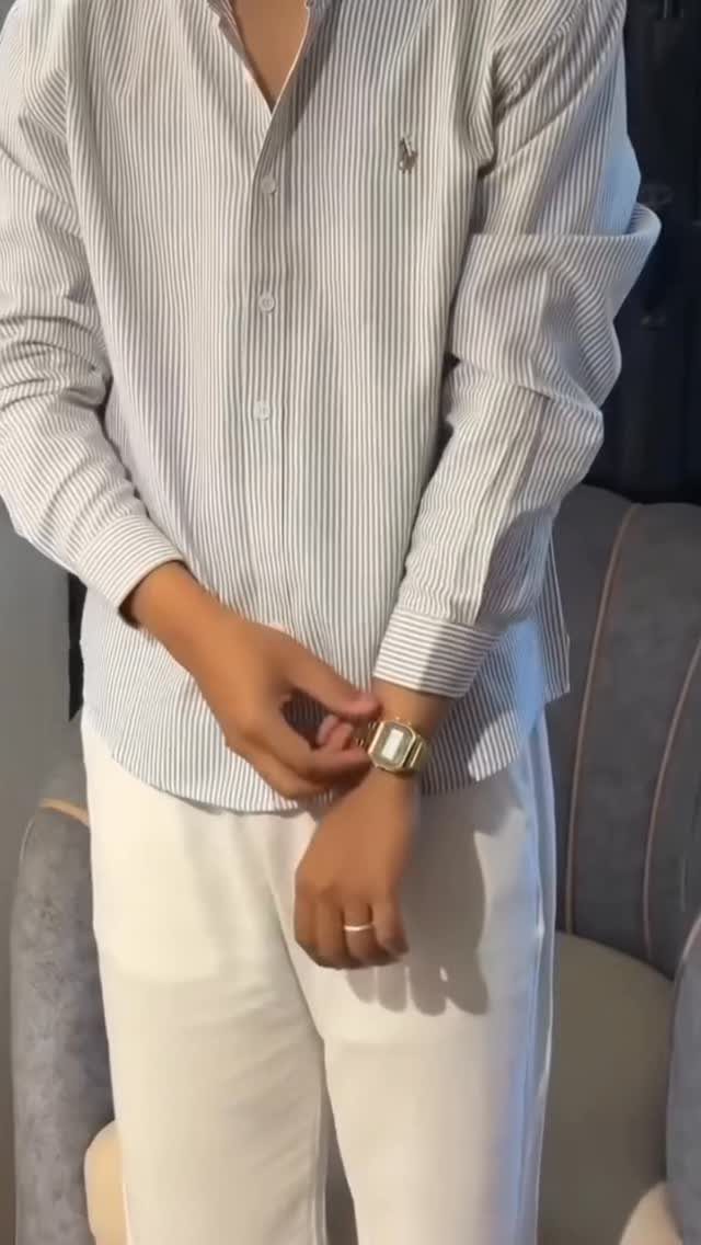 Man in a striped button-down shirt and white trousers adjusting a gold wristwatch.