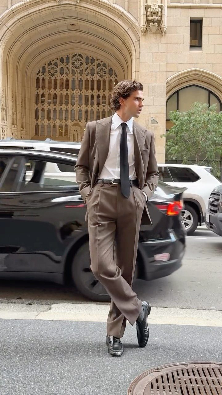 A man in a brown tailored suit and black tie posing on a city street.