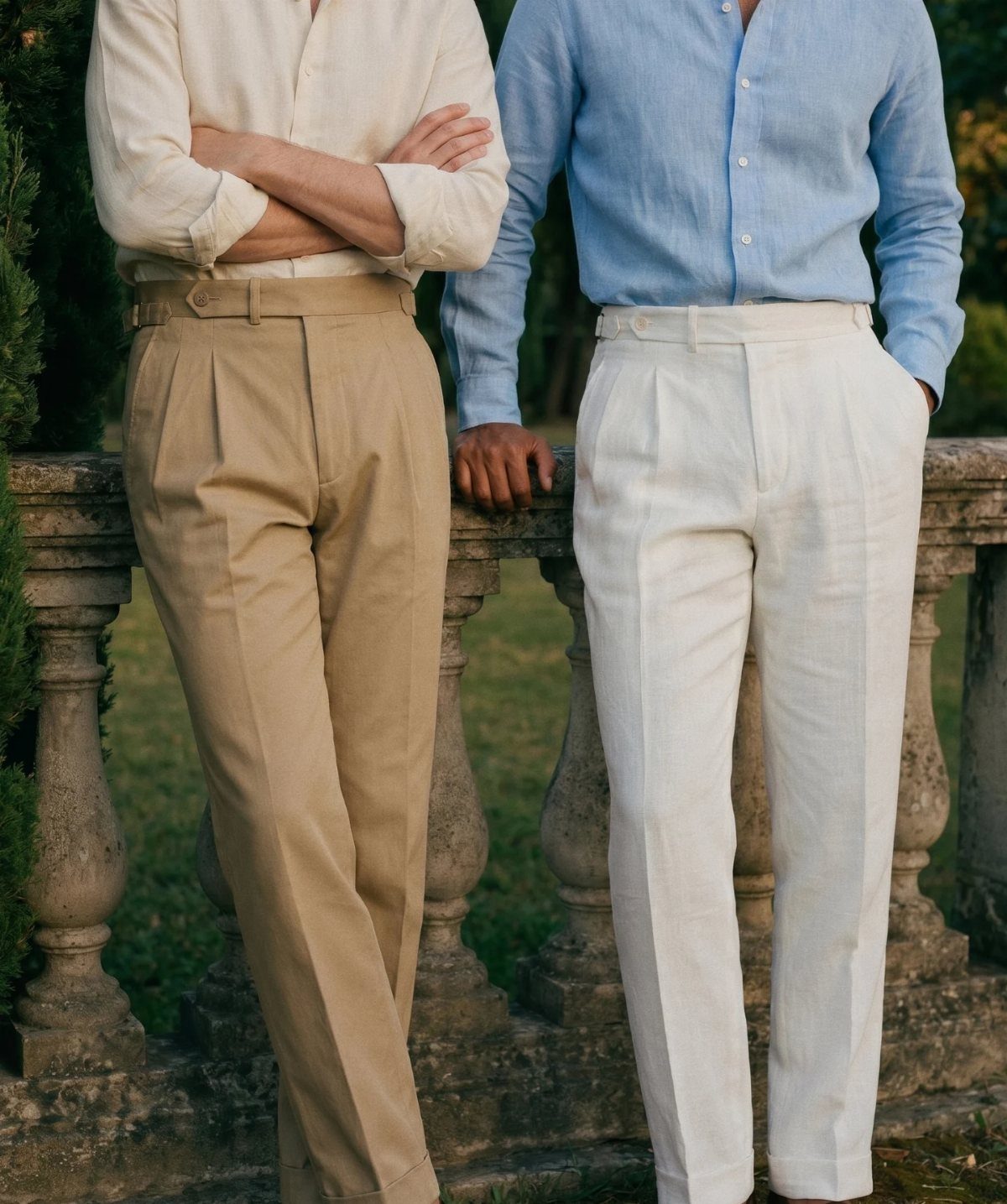 Two men wearing pleated linen trousers and shirts standing by a stone railing outdoors.
