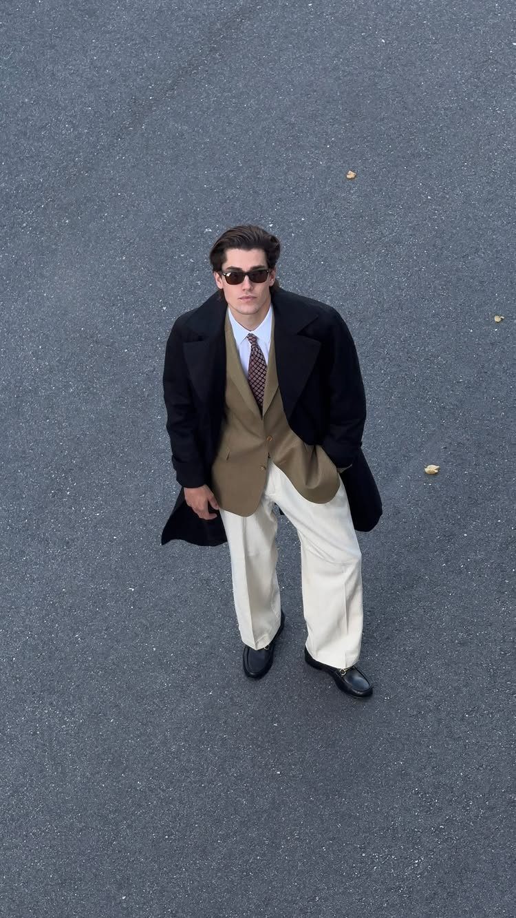 Man in a tan blazer, white shirt, tie, and cream trousers standing on asphalt.