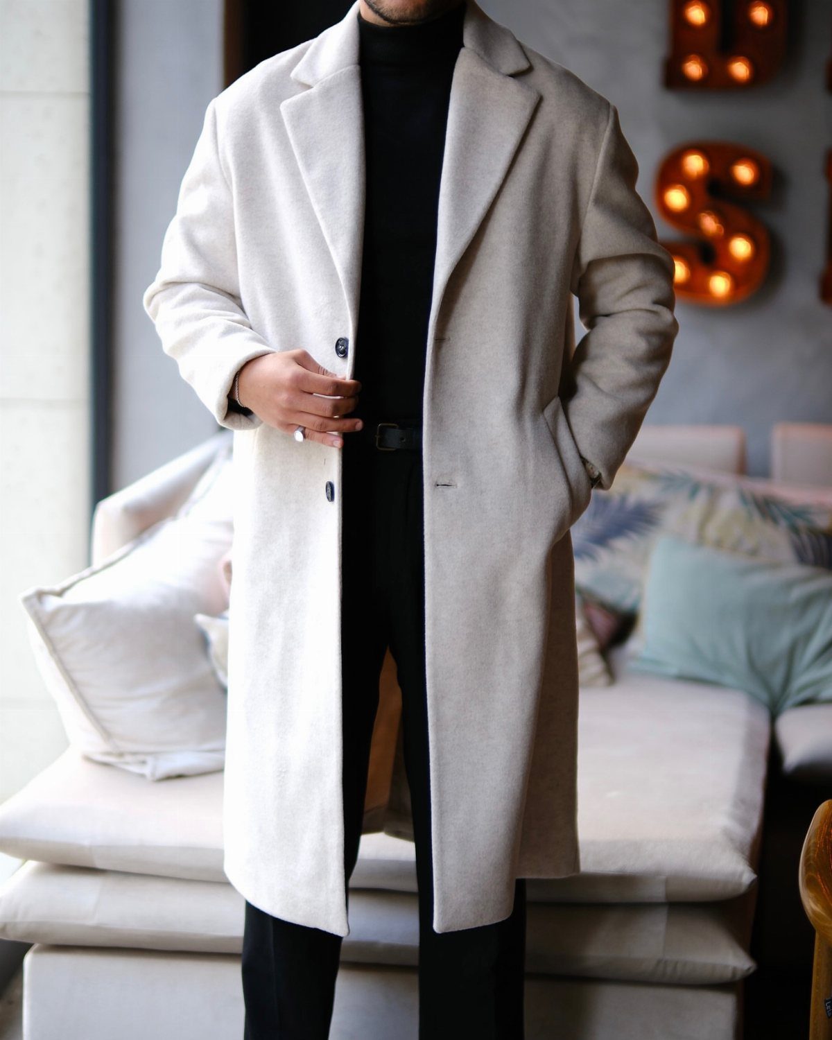 A man wearing a long cream wool overcoat, black turtleneck, and black trousers standing indoors.