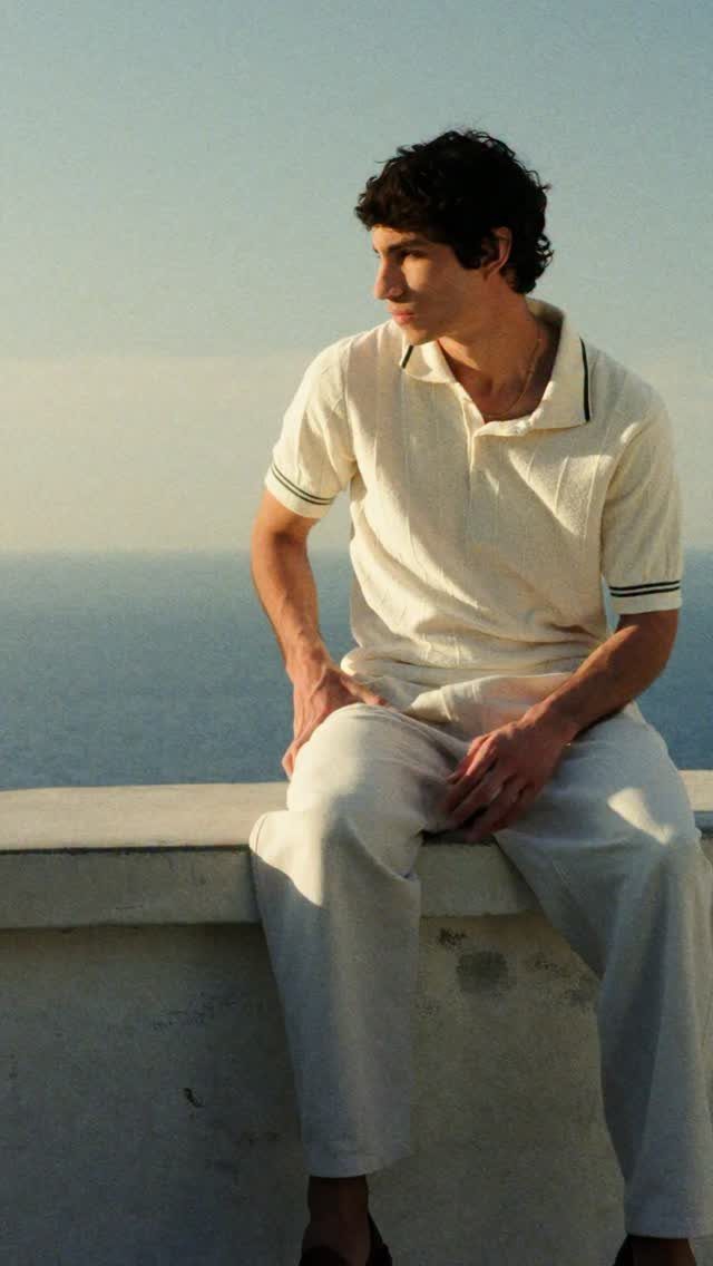 Man in a cream polo shirt and light trousers sitting by the sea at sunset.