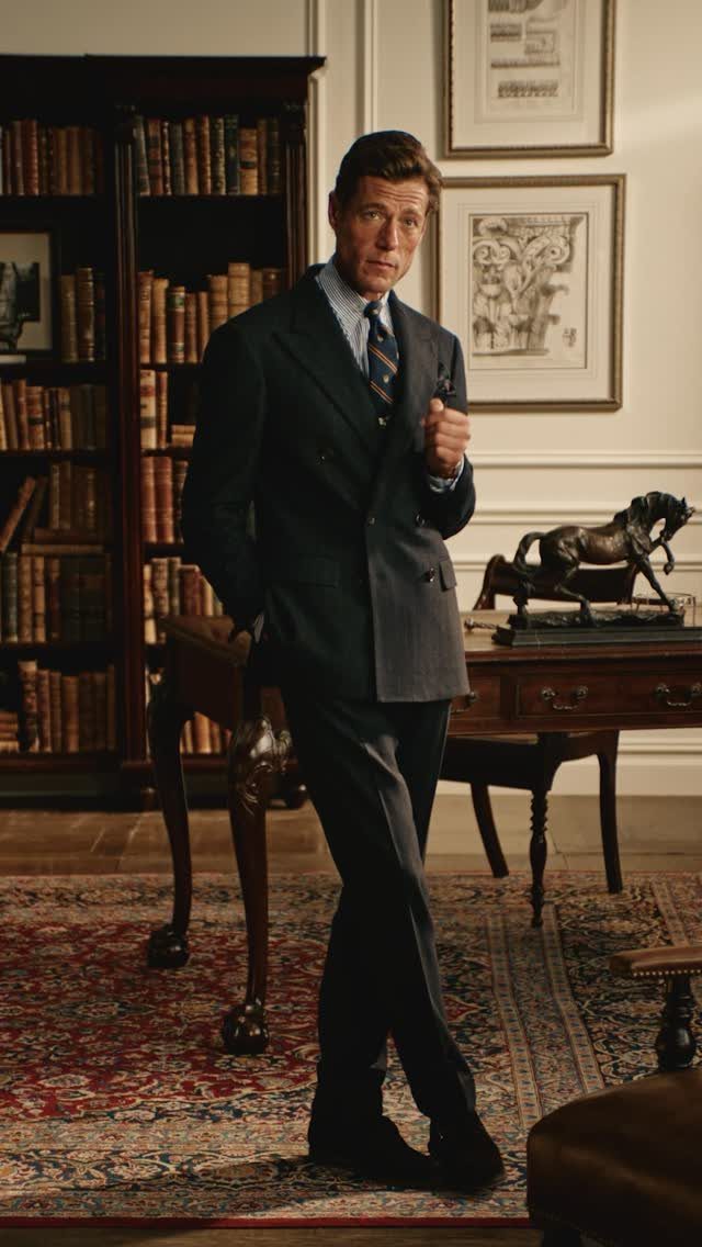 A man wearing a dark double-breasted tailored suit stands in a classic, wood-filled library study.