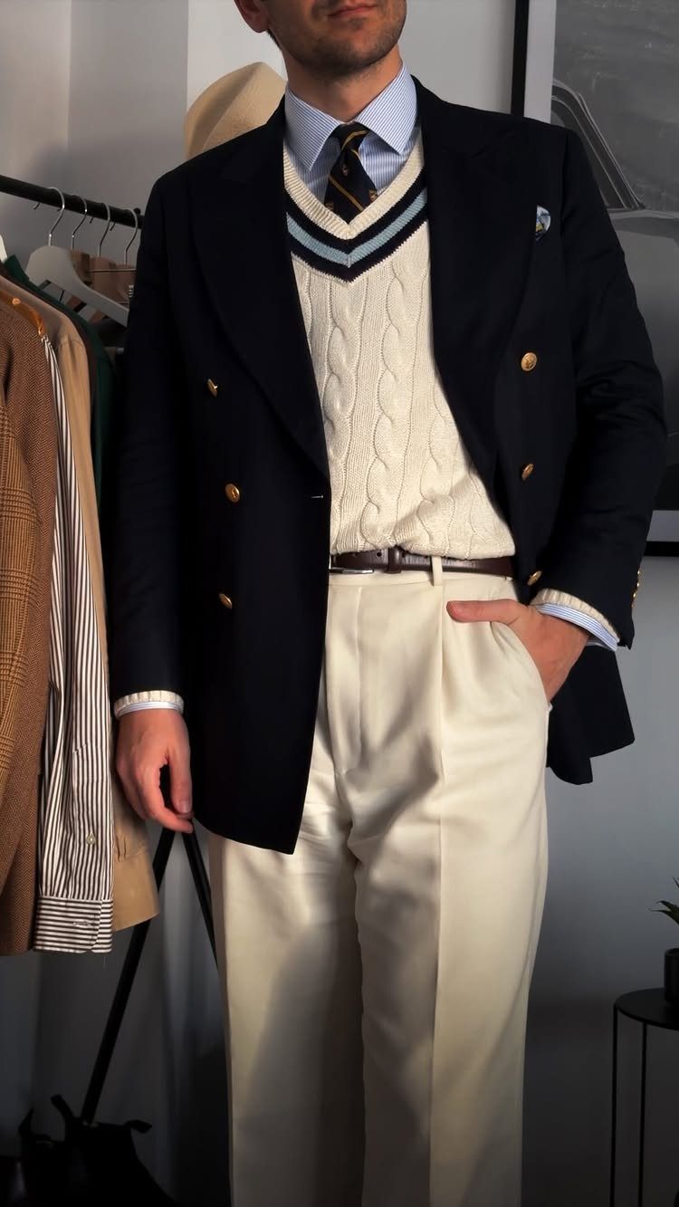 Man wearing a navy blazer, cream cable-knit sweater, and cream pleated tailored trousers.