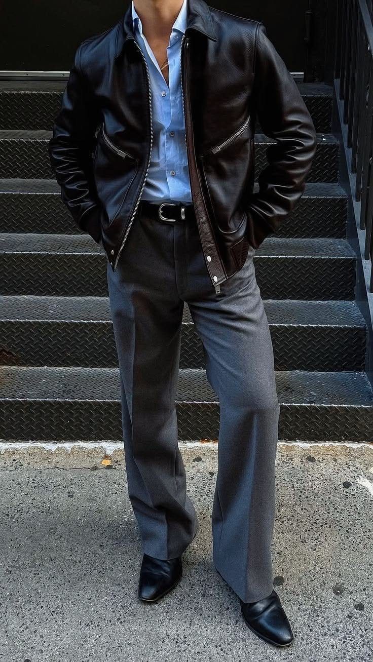 Man wearing a black leather jacket, blue shirt, and grey trousers standing on stairs.