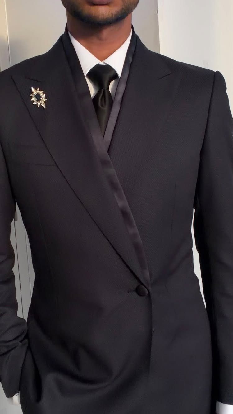 A man wearing a formal black tuxedo with a silver lapel brooch and black tie.