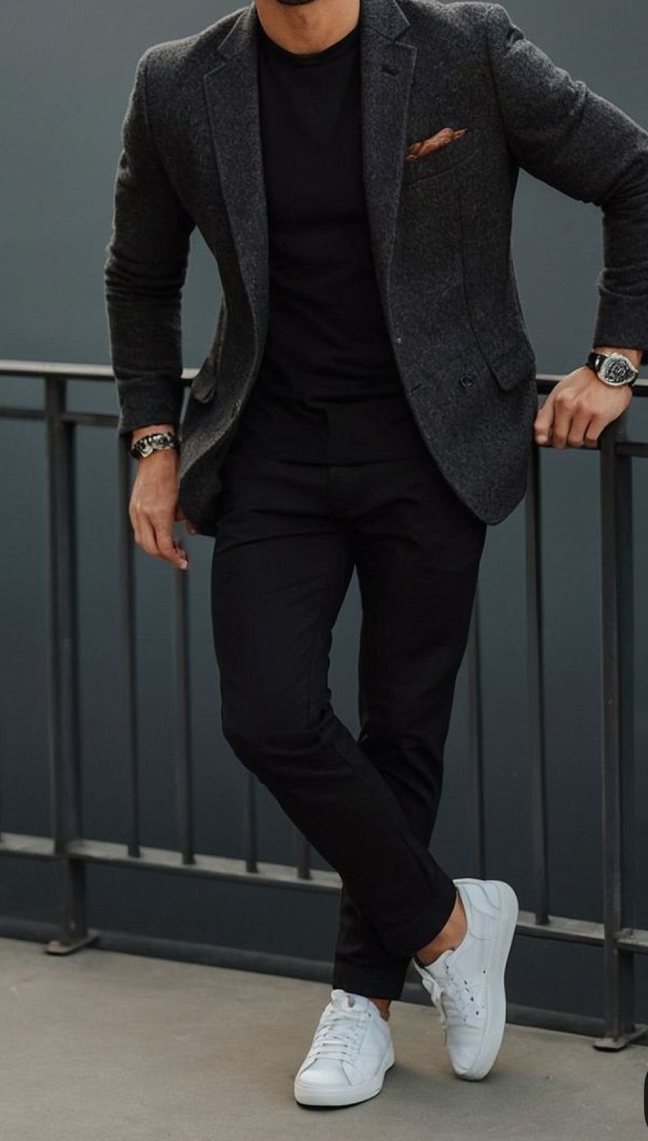 A man in a grey blazer, black t-shirt, black pants, and white sneakers leaning on a railing.