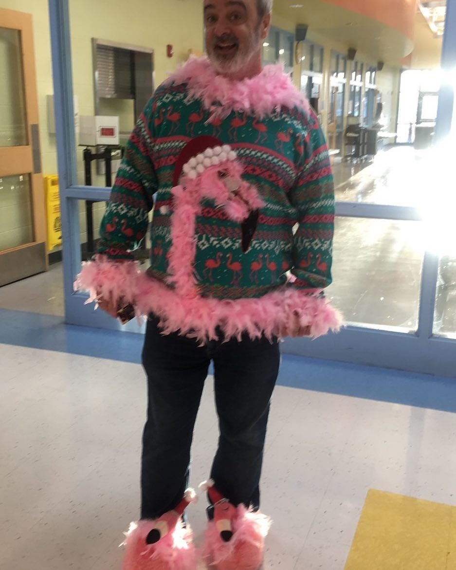 A man smiling in a green flamingo patterned sweater with pink feathers and matching slippers.