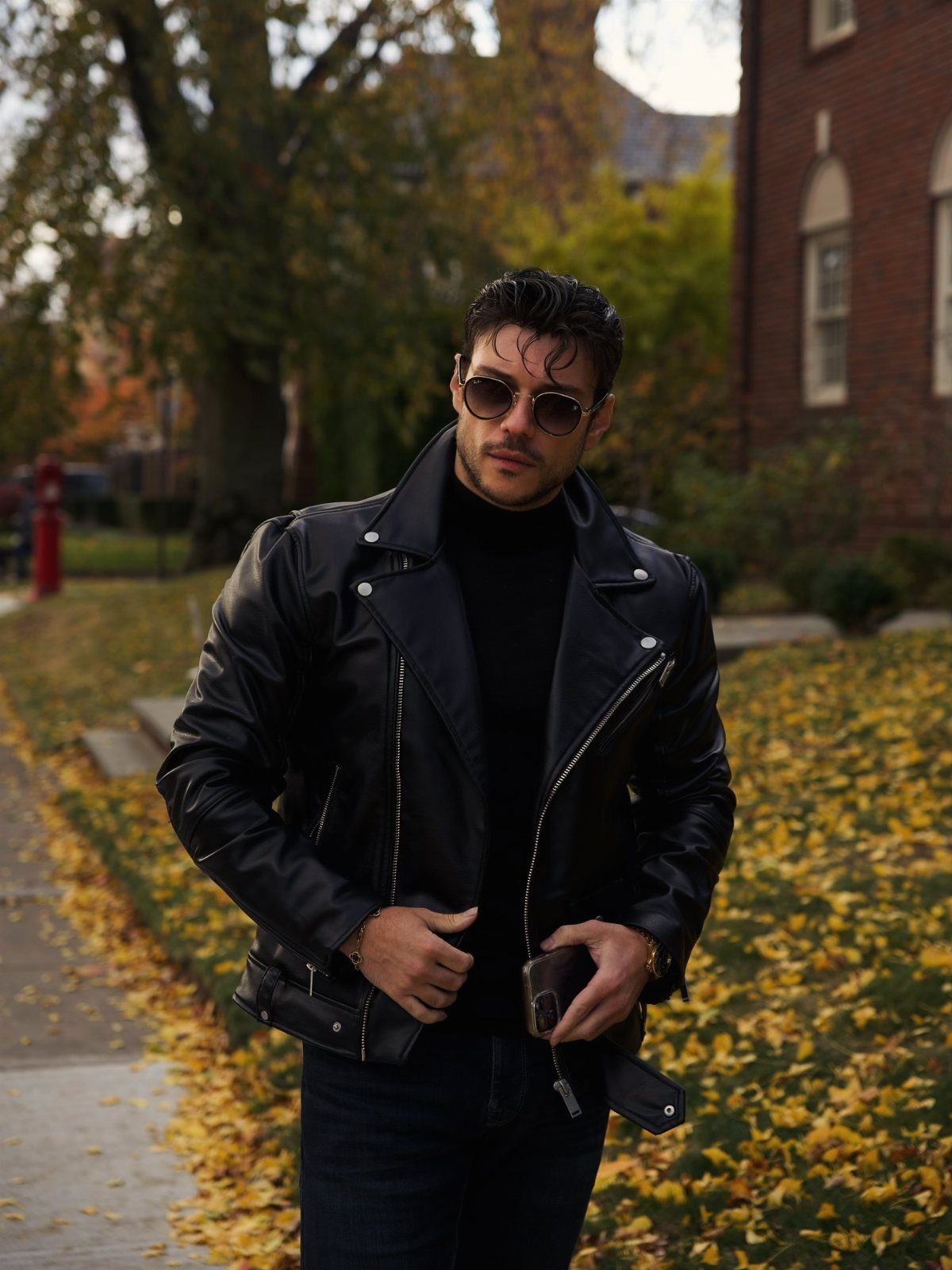Man in a black leather jacket, black turtleneck, and dark jeans standing in a park.