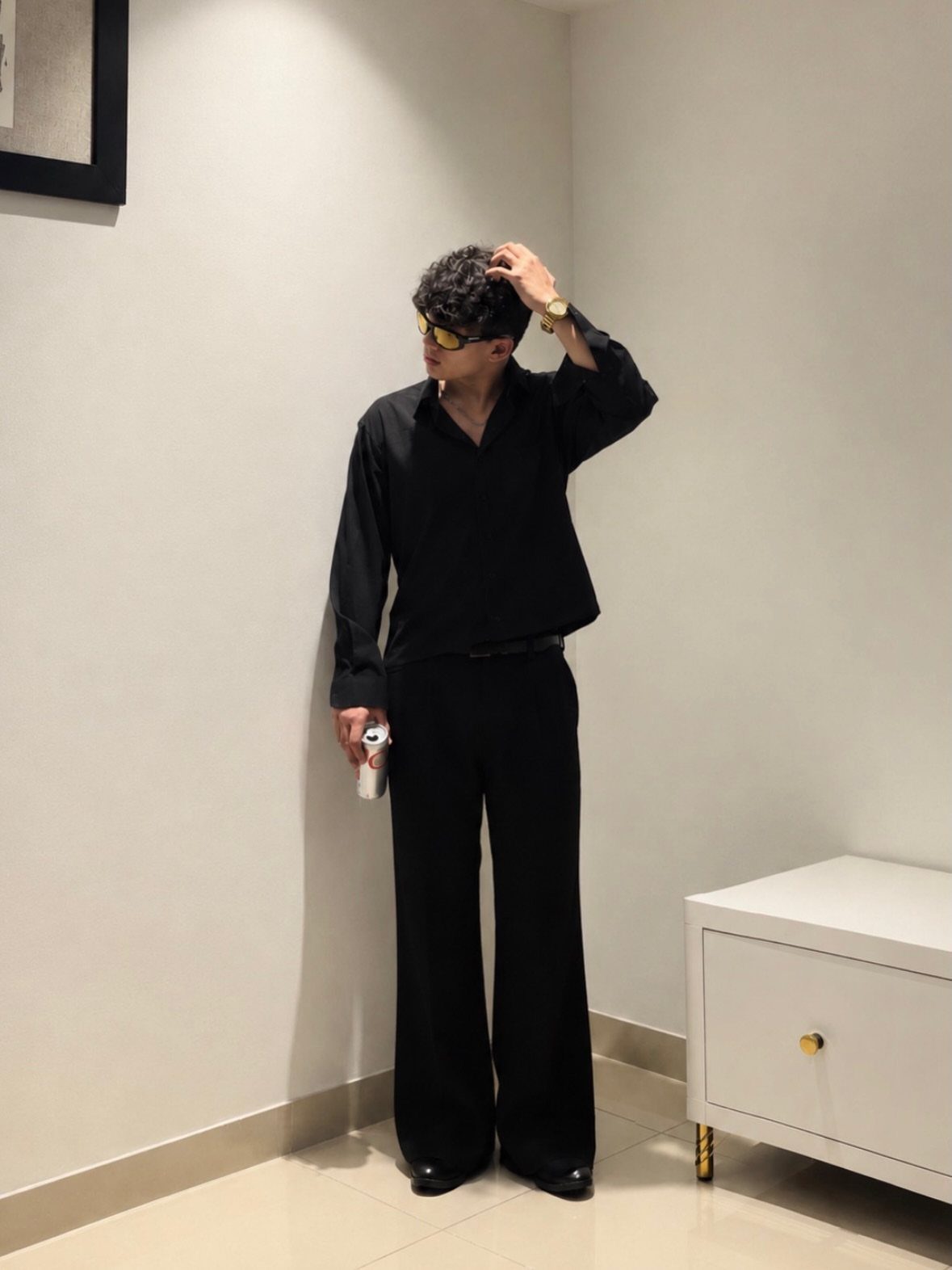 Man in an all-black outfit with wide-leg trousers and yellow sunglasses posing against a white wall.