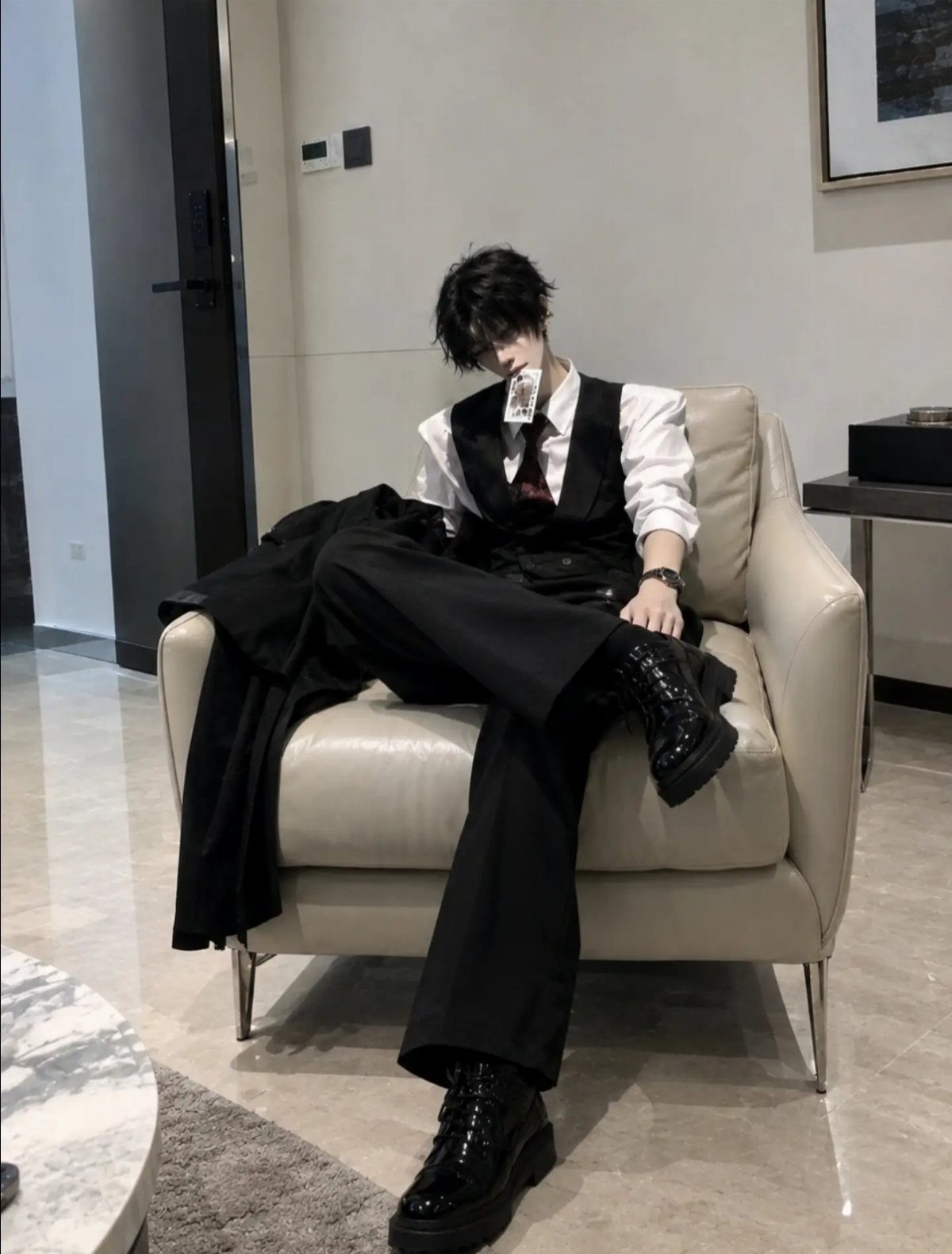 Man in black vest and trousers sitting on a white armchair wearing chunky black boots.