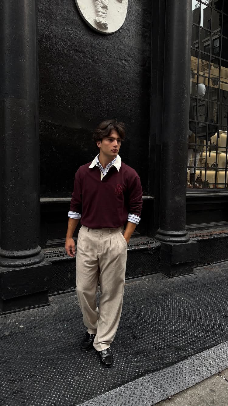 Man in burgundy sweater and beige pleated trousers standing against a dark building.