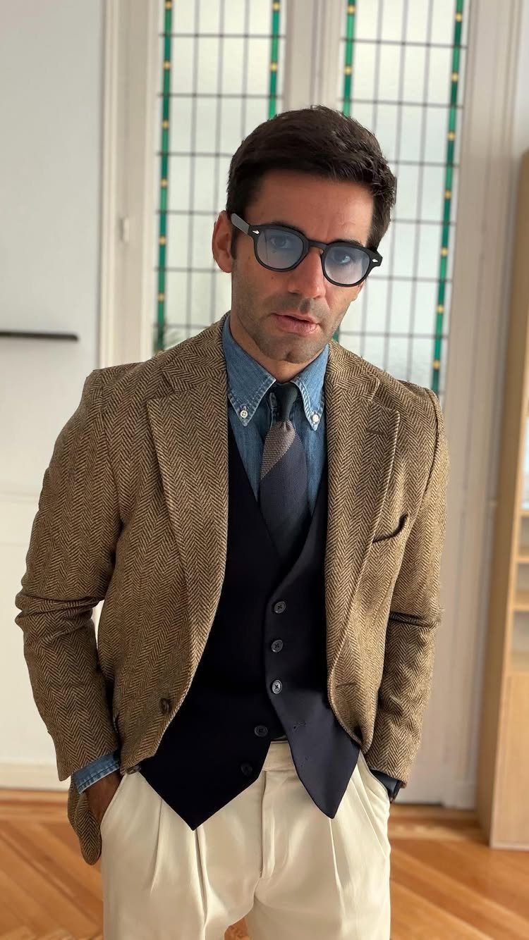 Man wearing a brown textured blazer, navy waistcoat, denim shirt, and cream trousers.