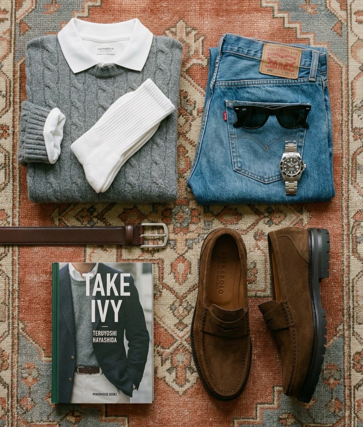 Brown suede loafers, denim jeans, a grey sweater, and a belt laid on a patterned rug.