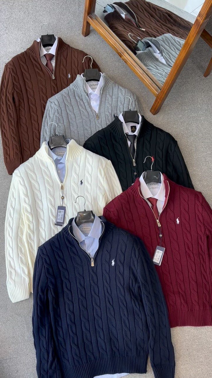 Multiple colorful cable-knit sweaters with collared shirts and ties hanging on a surface.