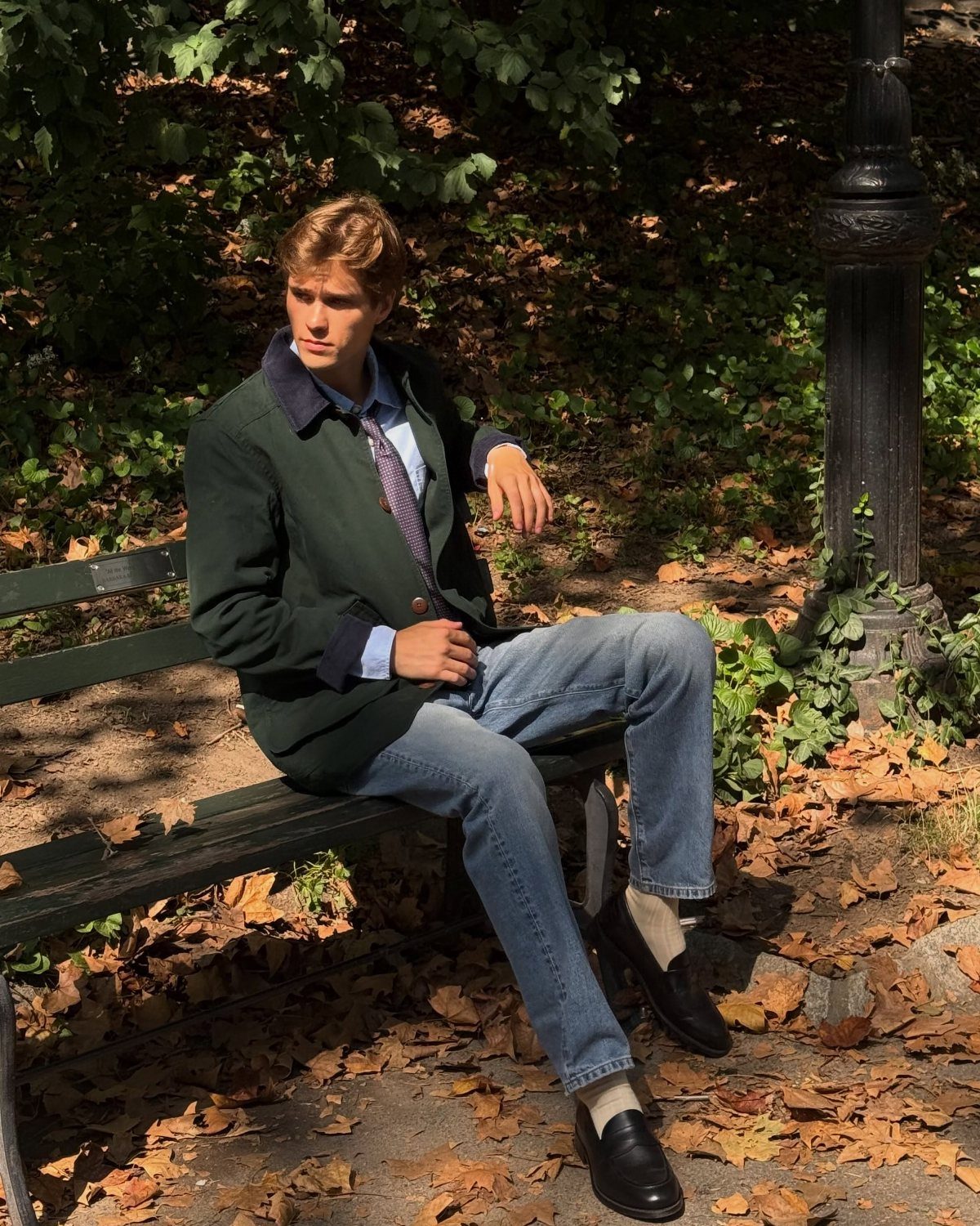 A young man wearing a dark green field jacket and blue jeans sits on a park bench.