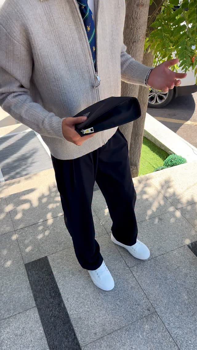 Man dressed in a beige cardigan, navy trousers, white sneakers, and a patterned tie holding a black clutch.