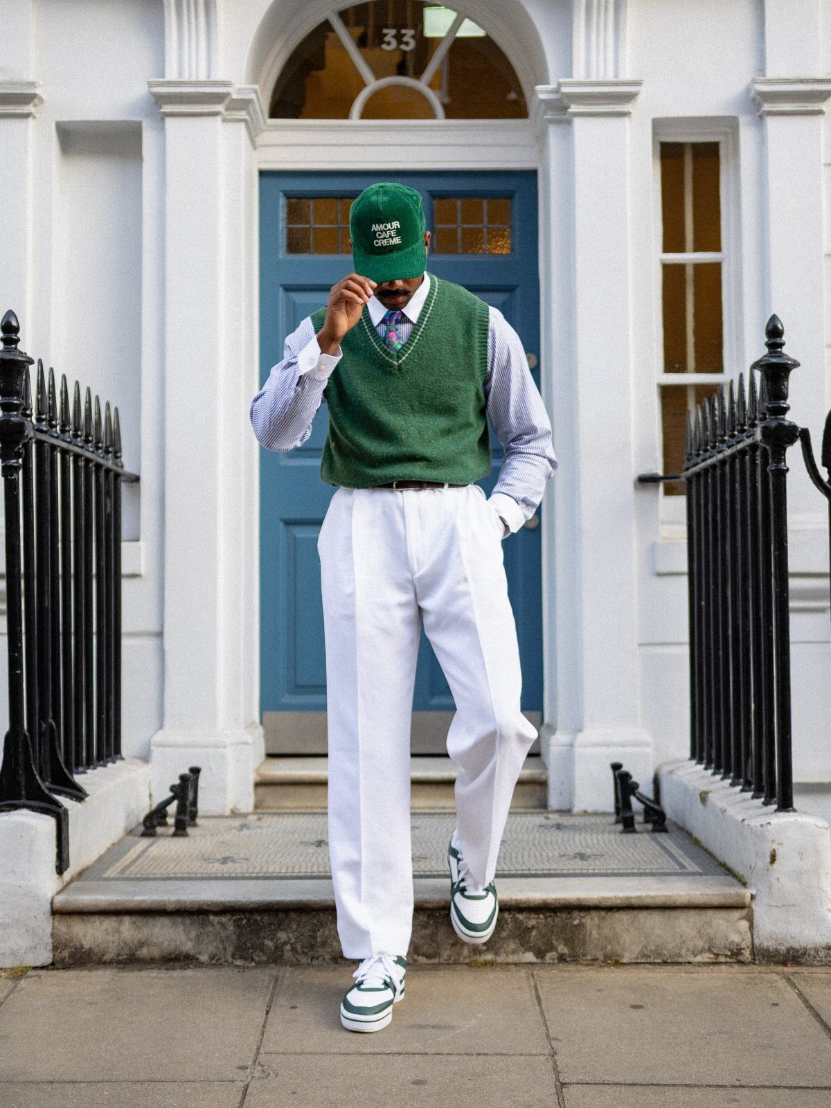 A man wearing a green sweater vest, white trousers, and a green cap walks down steps.