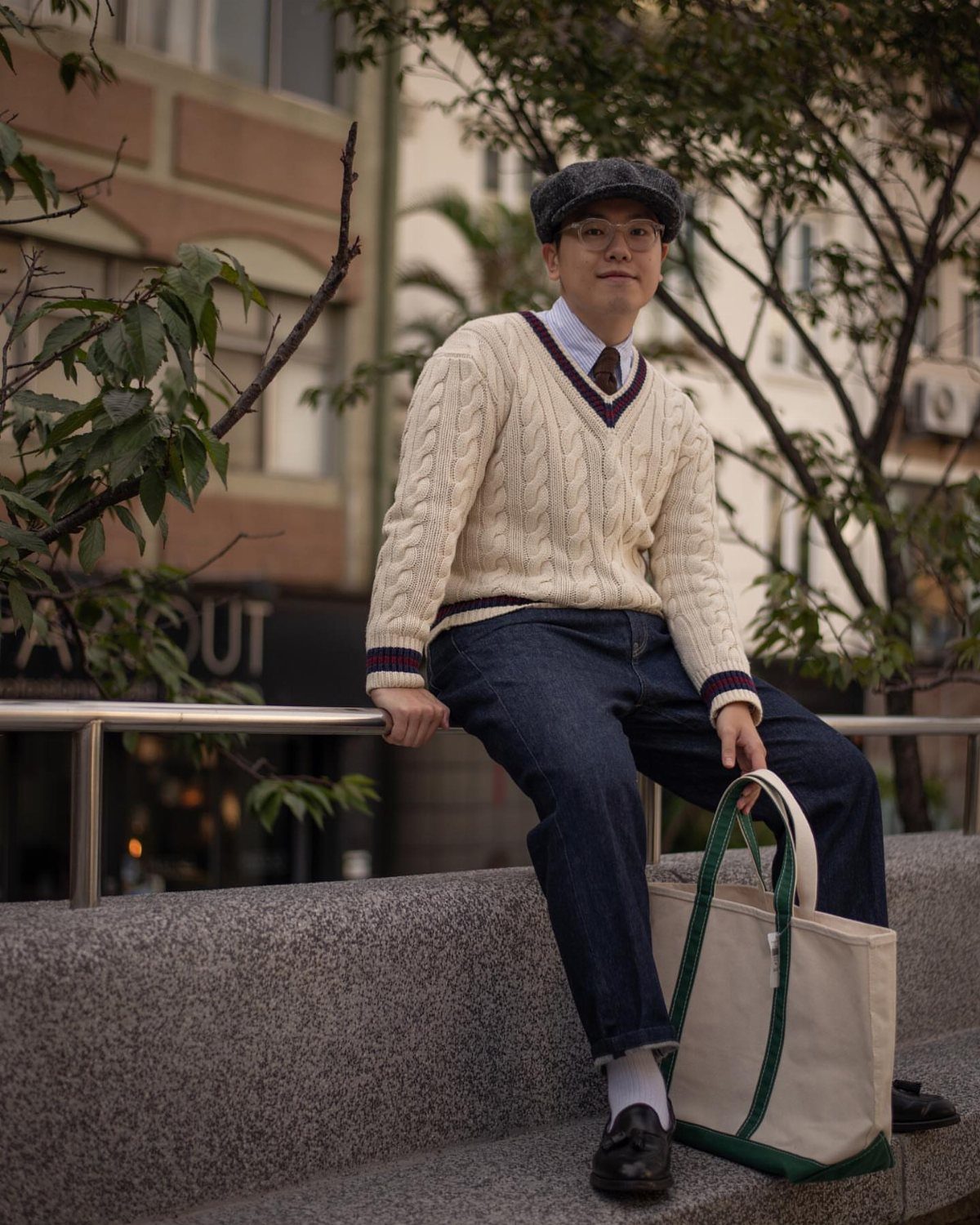 Man sitting on a stone ledge wearing a cream cable-knit sweater, dark jeans, and a flat cap.