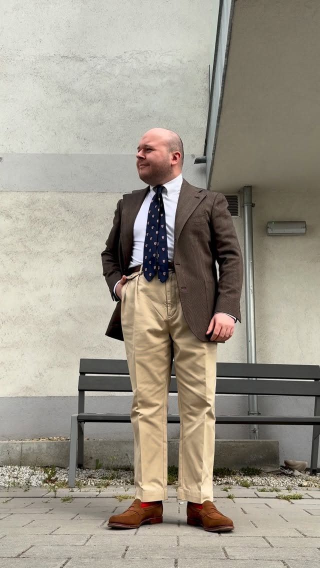 Man in a brown blazer, white shirt, navy tie, and tan chinos standing outdoors.
